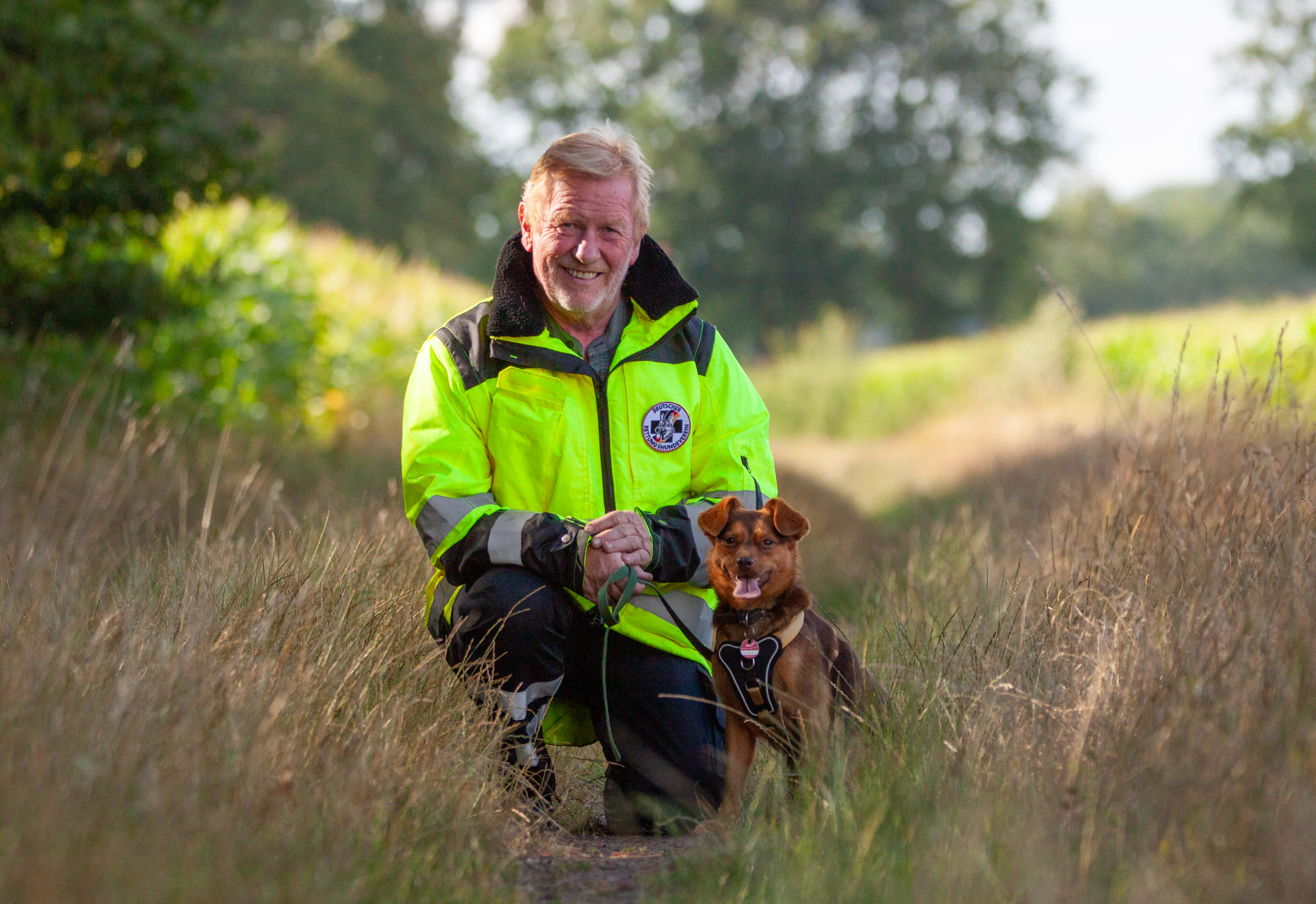 Dieter Granzow mit Mischling Emma Auf dem Bild sind ein Mann und ein kleiner Hund auf einem schmalen, naturbelassenen Weg zwischen hohen Gräsern abgebildet. Der Mann, Dieter Granzow, kniet freundlich lächelnd im Gras und hält eine grüne Leine in den Händen. Er trägt eine leuchtend gelbe Einsatzjacke mit reflektierenden Streifen und dem Logo des Deutschen Rettungshundevereins auf der Brust. Neben ihm sitzt seine Hündin Ema, eine kleine braune Mischlingshündin mit kurzen Beinen, spitz nach vorne gelappten Ohren und einem freundlichen Ausdruck. Sie trägt ein Hundegeschirr mit einem schwarzen Brustschild. Ema schaut aufmerksam in die Kamera und hat das Maul leicht geöffnet, sodass ihre Zunge sichtbar ist – sie wirkt fröhlich und wachsam. Der Hintergrund zeigt einen ruhigen Weg durch eine ländliche Landschaft mit grünen Feldern, Büschen und Bäumen. Das Bild strahlt Ruhe, Vertrautheit und Teamgeist zwischen Mensch und Hund aus.
