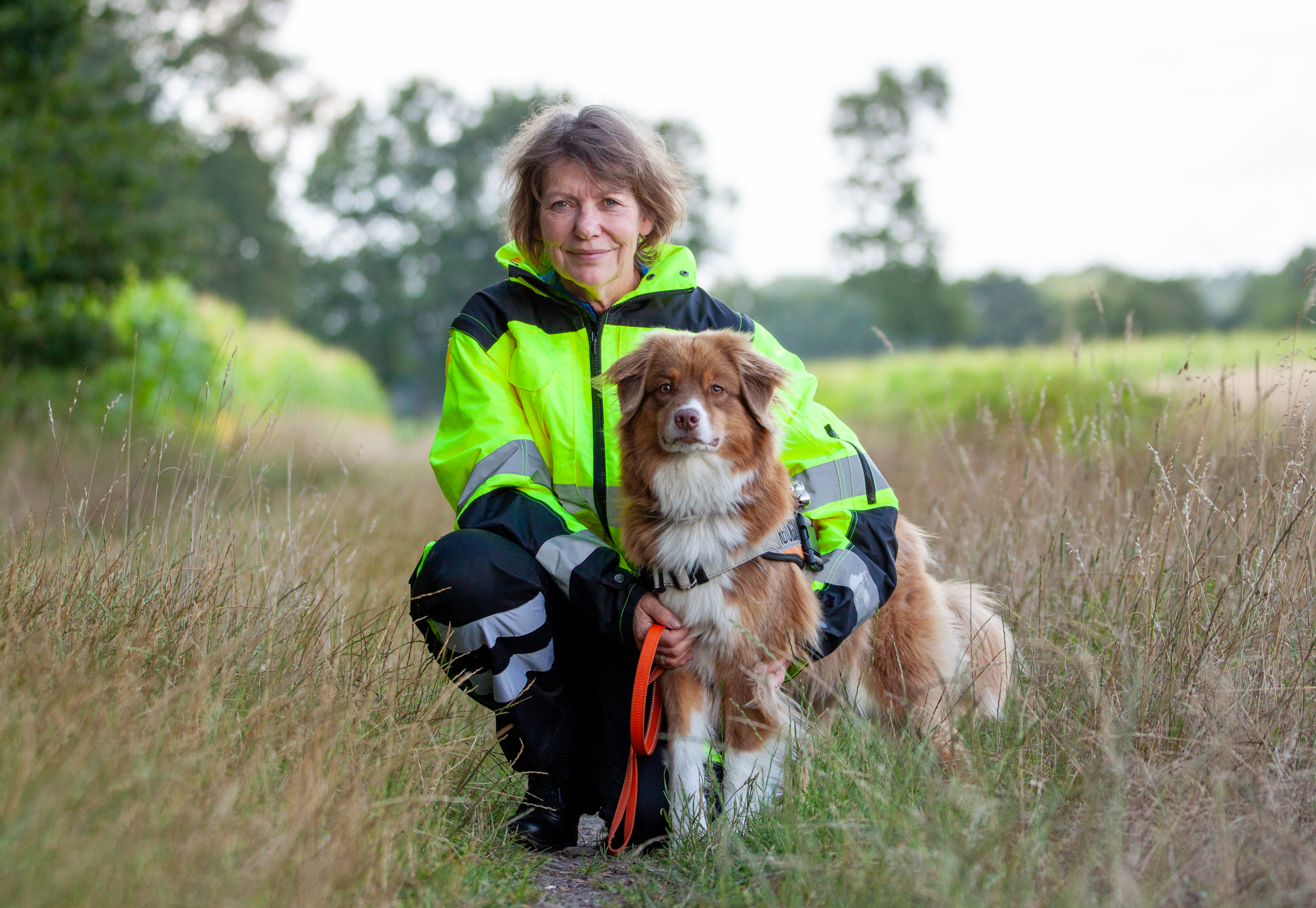 Martina Granzow mit Australian Shepherd Hund Elli Auf dem Bild sind eine Frau und ein Hund auf einem schmalen Naturweg zwischen hohen Gräsern zu sehen. Die Frau, Martina Granzow, kniet im Gras und hält ihre Hündin liebevoll an der Seite fest. Sie trägt eine auffällige, leuchtend gelbe Einsatzjacke mit reflektierenden Streifen und einem Abzeichen einer Rettungshundestaffel. Martina hat kurze, wellige, hellbraune Haare und lächelt ruhig in Richtung Kamera. Neben ihr sitzt ihre Australian Shepherd Hündin Elli. Elli hat ein auffälliges, mittellanges Fell in Braun-, Weiß- und Beigetönen. Sie trägt ein Hundegeschirr und eine orangefarbene Leine. Ihre aufmerksamen Augen sind direkt auf die Kamera gerichtet. Im Hintergrund sind Bäume und grüne Felder zu sehen. Die Szene wirkt ruhig, verbunden mit der Natur, und strahlt Vertrauen und Teamgeist zwischen Mensch und Hund aus.