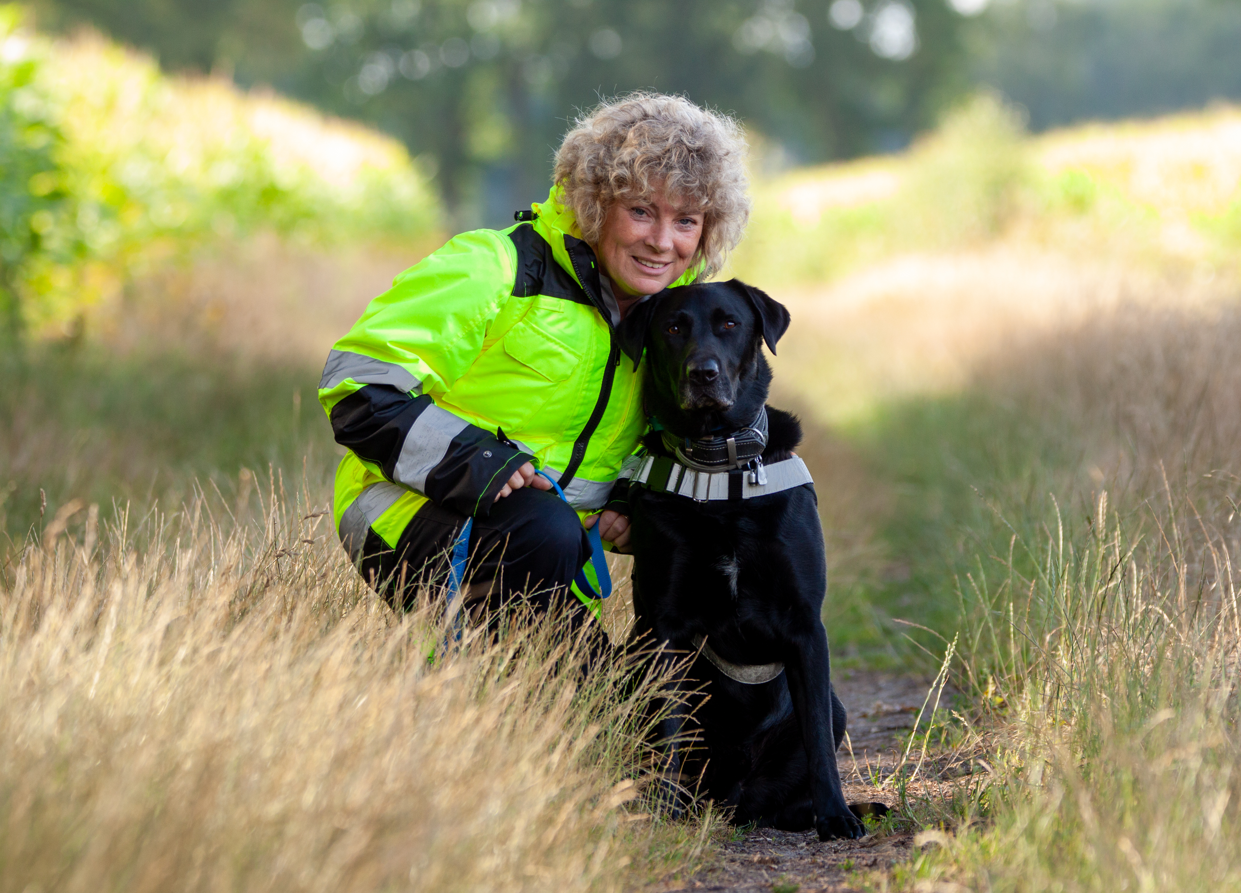 Tanja Goldmann und Labrador Cooper Das Bild zeigt Tanja Goldmann mit lockigen, blonden Haaren, die auf einem schmalen, naturbelassenen Weg kniet. Neben ihr sitzt ihr schwarzer Hund namens Cooper. Tanja trägt eine auffällige, neongelbe Jacke mit reflektierenden Streifen – typisch für Einsatzkleidung. Sie lächelt freundlich und hat eine Hand auf dem Rücken ihres Hundes gelegt. Cooper – ein Labrador Retriever – trägt ein gut sichtbares, reflektierendes Geschirr, wie es bei Rettungshunden üblich ist. Beide blicken direkt in die Kamera. Die Szene spielt sich bei Tageslicht in einer sommerlichen Landschaft ab, mit hohem Gras rechts und links des Weges sowie verschwommenen Bäumen im Hintergrund. Die Atmosphäre wirkt ruhig und harmonisch.