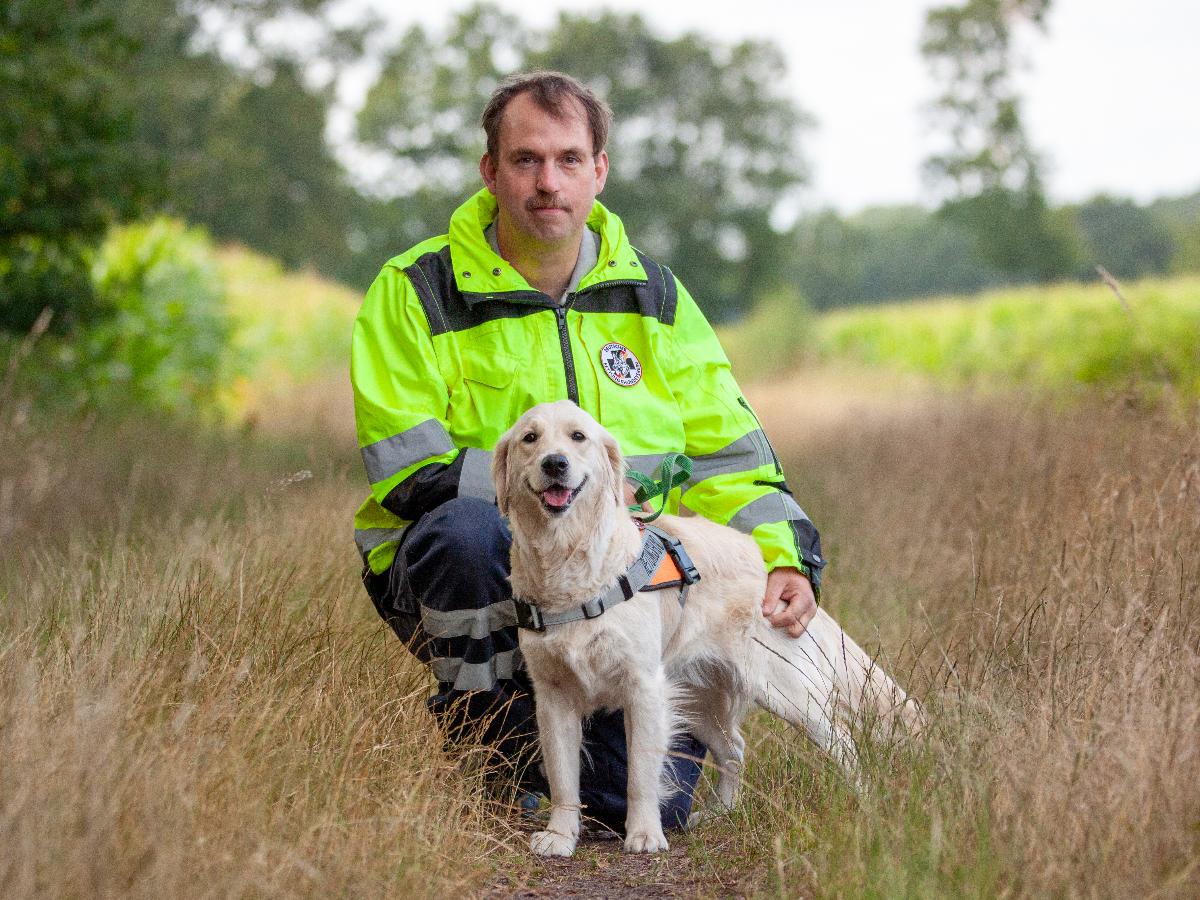 Andreas Scheper und Golden Retriever Tala Auf dem Foto ist Andreas Scheper in auffälliger Einsatzkleidung zusammen mit seinem Hund auf einem schmalen, naturbelassenen Weg zu sehen. Andreas trägt eine leuchtend gelbe Jacke mit reflektierenden Streifen und kniet im Gras. Auf seiner Jacke ist ein rundes Abzeichen mit dem Logo vom Deutschen Rettungshundeverein zu erkennen. Neben ihm steht seine freundliche, mittelgroße, hellbeige bis creamfarbige Golden Retriever Dame „Tala“. Tala trägt ein Geschirr mit auffälligen orange Farben, das auf ihre Funktion als Rettungshund hinweist. Beide schauen direkt in die Kamera. Im Hintergrund sind Bäume und ein grünes Feld zu sehen, es ist ein heller, leicht bewölkter Tag. Die Szene wirkt ruhig und harmonisch.