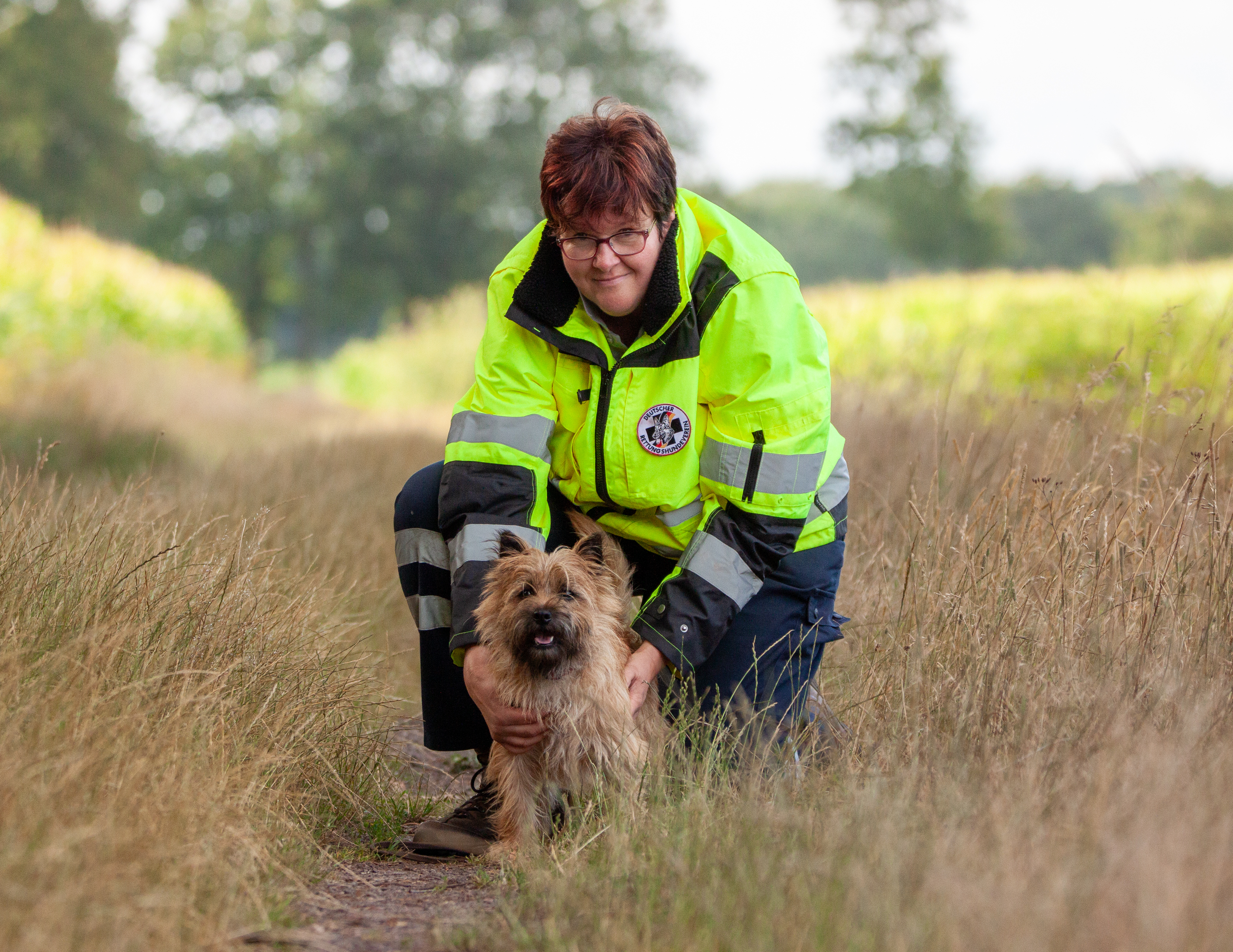 Katrin Werdin mit Cairn Terrier Teddy Auf dem Bild sind eine Frau und ein kleiner Hund auf einem naturbelassenen Weg in einer ländlichen Umgebung zu sehen. Die Frau, Katrin Werdin, kniet im Gras und hält ihren Hund liebevoll an beiden Seiten. Sie trägt eine auffällige, leuchtend gelbe Einsatzjacke mit reflektierenden Streifen und einem runden Abzeichen einer Rettungshundestaffel. Katrin hat kurze, rotbraune Haare und trägt eine Brille. Ihr Blick ist freundlich und sie schaut in Richtung Kamera. Vor ihr sitzt ihr Hund Teddy, ein kleiner Cairn Terrier mit zotteligem, beigefarbenem Fell und dunkler Schnauze. Teddy schaut ebenfalls aufmerksam in die Kamera, sein Maul ist leicht geöffnet, sodass es aussieht, als würde er freundlich „lächeln“. Im Hintergrund erstreckt sich ein Weg durch hohes Gras, flankiert von Bäumen und grünen Feldern. Die Szene wirkt ruhig, naturverbunden und zeigt ein harmonisches Team aus Mensch und Hund.